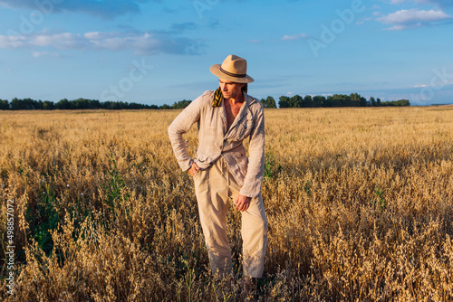 Rural Countryside. Tall handsome man dressed in a coarse linen suit and hat standing at golden oat field © Smile