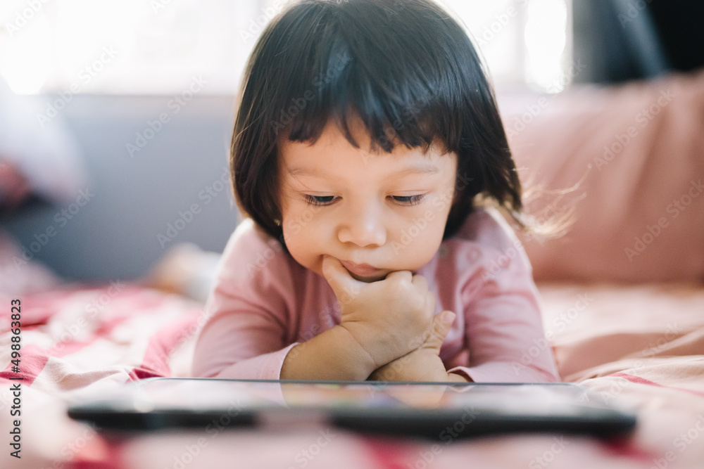 A little girl in a pink shirt lay intently playing the tablet on her bed. self-learning technology education concept A little girl in a pink shirt lay intently playing the tablet on her bed. self-learning technology education concept
