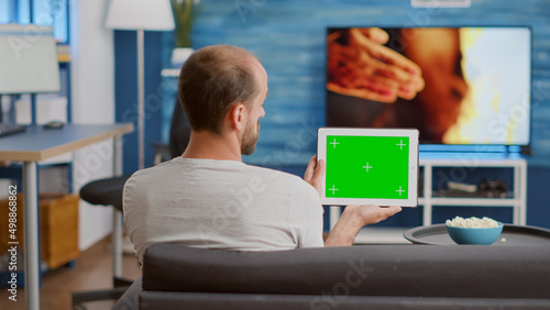 Back view of man holding digital tablet with green screen watching social media video content sitting on sofa. Person looking at touchscreen device with chroma key looking at influencer vlog.