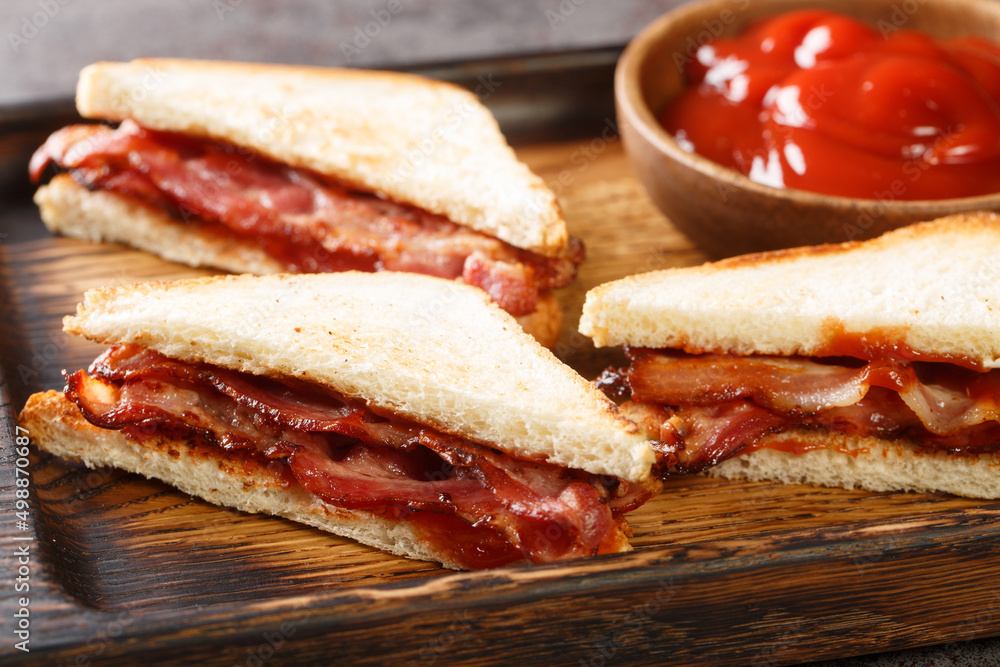Bacon butty is a British sandwich consisting of crispy bacon, butter, and sauce closeup in the wooden tray on the table. Horizontal Bacon butty is a British sandwich consisting of crispy bacon, butter, and sauce closeup in the wooden tray on the table. Horizontal