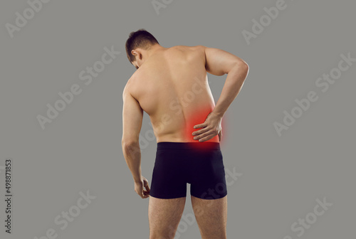 Backside of young man suffering from pain in right side. Patient in underwear standing isolated on grey background and holding hand on his inflamed lower back, rear view from behind. Backache concept