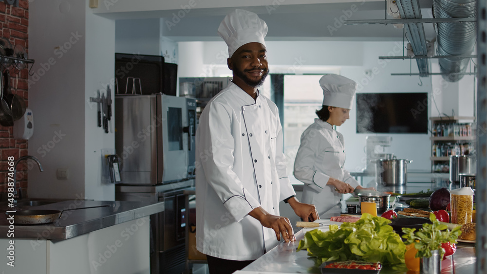 Portrait of african american chef cooking gourmet meal in busy restaurant kitchen, preparing food menu. Professional cook in uniform making cuisine dish with fresh ingredients, culinary recipe.