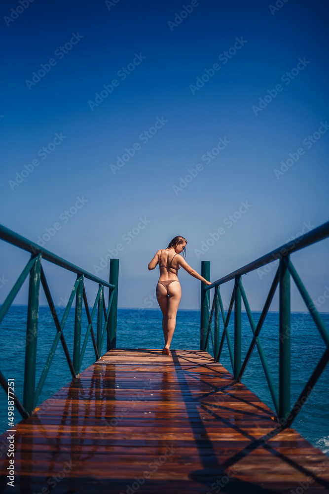 beautiful young woman in a brown swimsuit walks on the pier by the beach on the sea on a summer sunny day