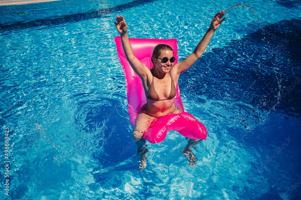 Sexy woman in sunglasses resting and sunbathing on a pink mattress in the pool. Young woman in beige bikini swimsuit floating on inflatable pink mattress