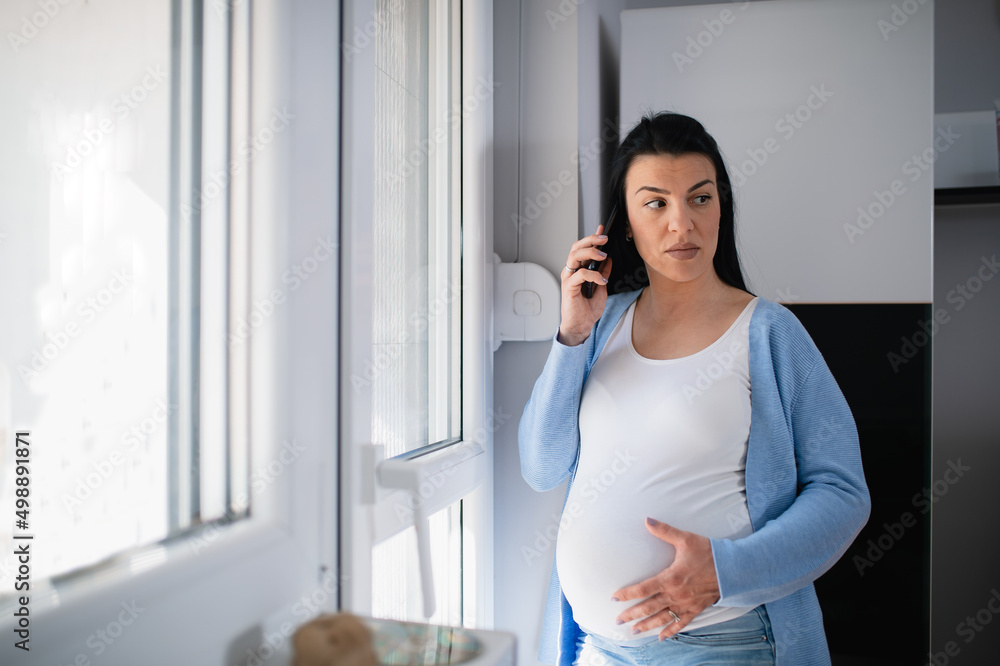 Unhappy pregnant caucasian brunette uses mobile phone and holds it on her ear while she seriously looks at her left side somewhere. She holds her big tummy with her left hand. Woman stands near door. Unhappy pregnant caucasian brunette uses mobile phone and holds it on her ear while she seriously looks at her left side somewhere. She holds her big tummy with her left hand. Woman stands near door.