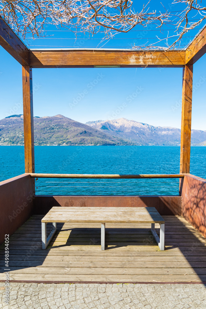Viewpoint on the blue rippled waters of Lake Maggiore, in the village of Luino, Lombardy, Italy. Italian alps with blue sky on the background. Wooden bench in the foreground.