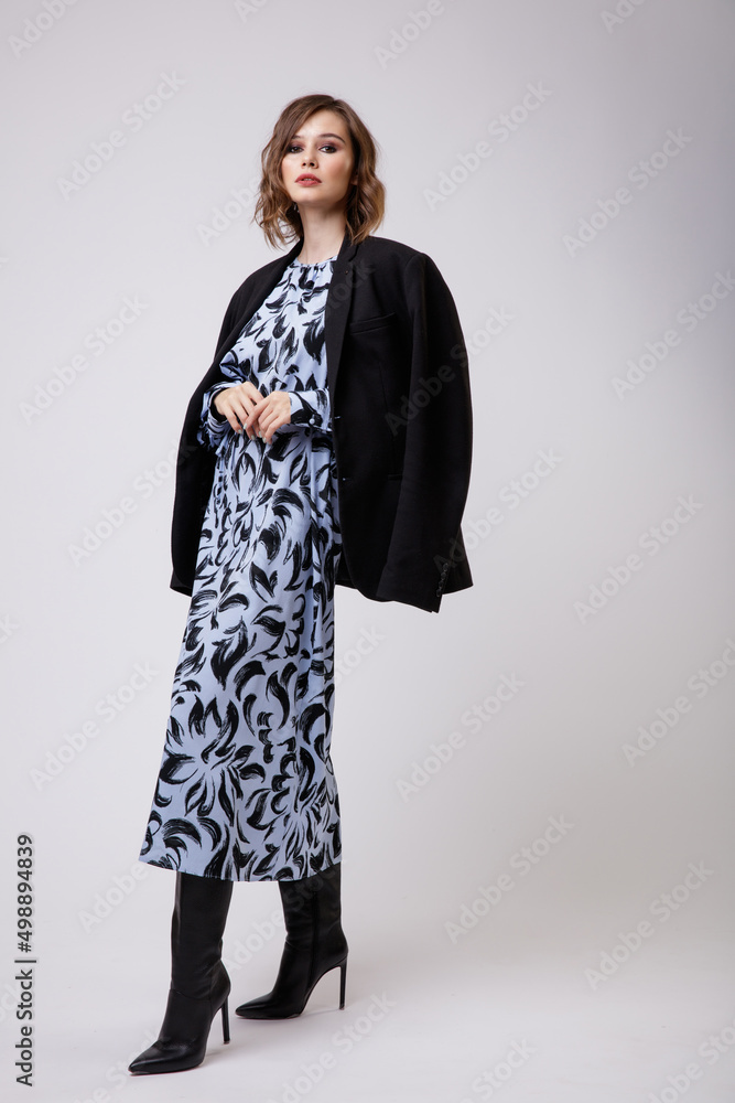 High fashion photo of a beautiful elegant young woman in a pretty long blue dress with floral pattern, black jacket, boots posing on white background. Slim figure, hairstyle, studio shot.