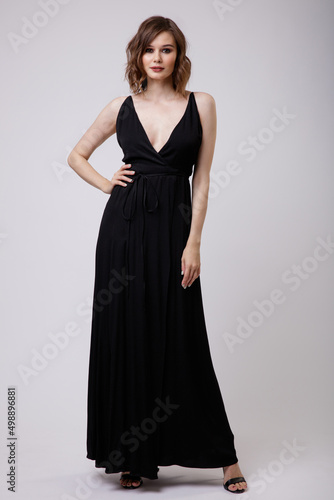High fashion photo of a beautiful elegant young woman in a pretty black evening party dress with a deep neckline posing on white background. Slim figure, hairstyle, studio shot. 