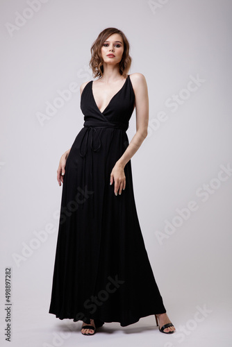 High fashion photo of a beautiful elegant young woman in a pretty black evening party dress with a deep neckline posing on white background. Slim figure, hairstyle, studio shot. 