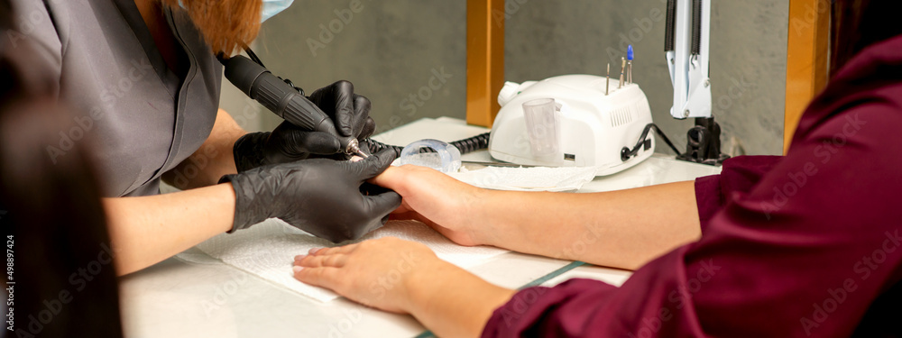 Obraz premium Manicure master wearing protective mask uses electric nail file machine in a nail salon, close up