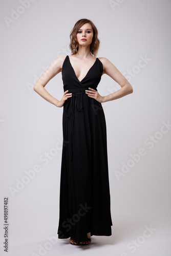 High fashion photo of a beautiful elegant young woman in a pretty black evening party dress with a deep neckline posing on white background. Slim figure, hairstyle, studio shot. 