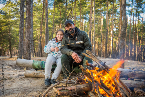 In autumn, on a bright sunny day in a pine forest, a father and daughter lit a fire and roast marshmallows on fire.