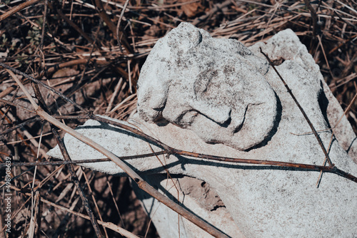A piece of toppled stone crucifix sculpture on the ground at abandoned ancient cemetery. Broken tombstone at the old churchyard. Cemetery vandalism