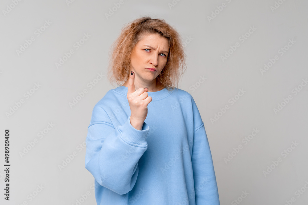 Serious middle age woman with curly blonde hair shakes her finger threateningly, scolding or telling off someone, standing in trendy blue sweatshirt over light grey background Serious middle age woman with curly blonde hair shakes her finger threateningly, scolding or telling off someone, standing in trendy blue sweatshirt over light grey background
