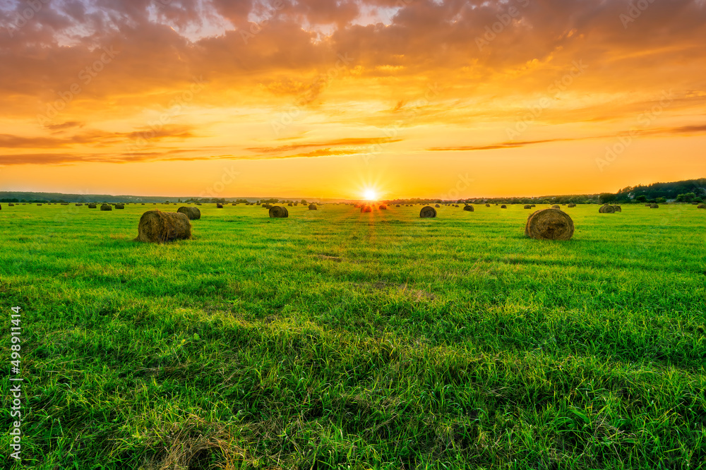 Scenic view at picturesque burning sunset in a green shiny field with hay stacks, bright cloudy sky , trees and golden sun rays, summer valley landscape