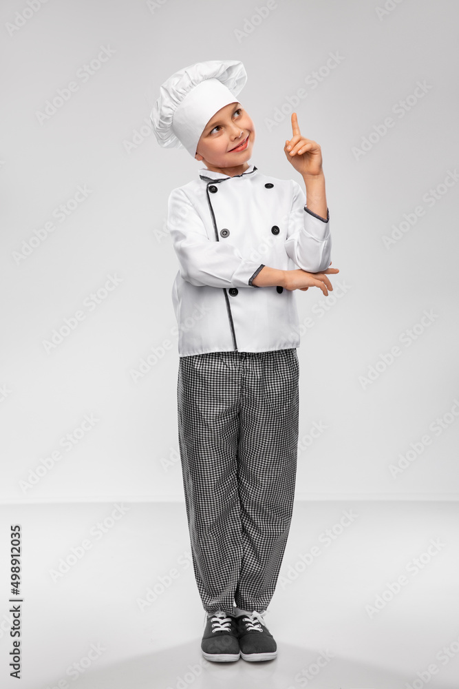 cooking, culinary and profession concept - happy smiling little boy in chef's toque and jacket pointing finger up over grey background