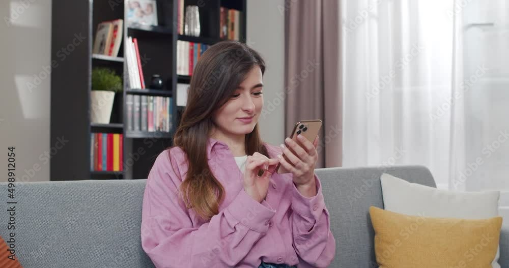 Smiled pretty Caucasian young female holding mobile phone and tapping on screen. At cozy home. Beautiful woman texting sms on smartphone. Answering in chat in social media. Chatting by messages. Smiled pretty Caucasian young female holding mobile phone and tapping on screen. At cozy home. Beautiful woman texting sms on smartphone. Answering in chat in social media. Chatting by messages.