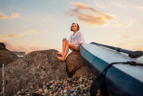 Summer activity vacations. Pre-school girl sitting with sup board at big beach rocks. Bottom view. Sunset sky at the background