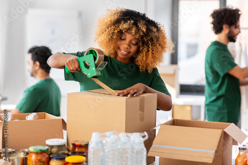 charity, donation and volunteering concept - happy smiling woman with adhesive tape packing food in boxes over international group of volunteers at distribution or refugee assistance center photo