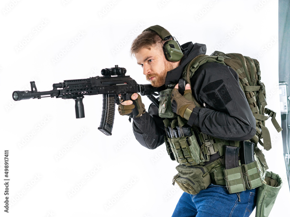 airsoft player in full gear with GG RK74 fire series guns. a man in headphones, a bulletproof vest, with a backpack and a belt, aims his machine gun to the side. profile. White background.