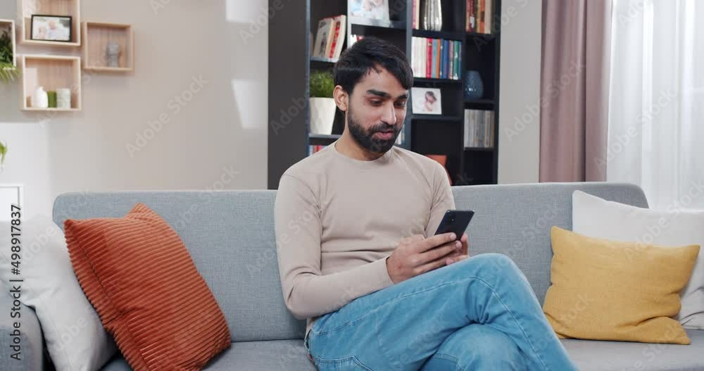 Happy Arabian young man sitting on sofa and chatting by messages on smartphone. At home. Smiled handsome male using mobile phone for texting sms. Tapping, typing and messaging on cellphone. Happy Arabian young man sitting on sofa and chatting by messages on smartphone. At home. Smiled handsome male using mobile phone for texting sms. Tapping, typing and messaging on cellphone.