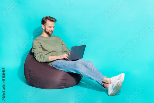 Full size photo of young blond guy type laptop wear sweater jeans shoes isolated on turquoise background