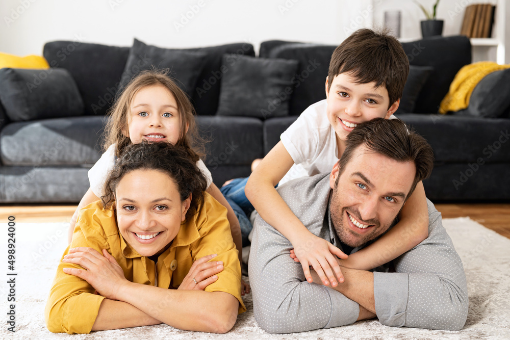 Happy multiracial family of four laying at the floor in cozy living room, parents and kids laughing while embracing with each other. Family feeling happy during the weekend