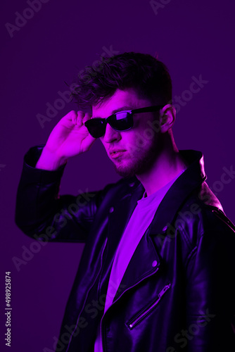 Young man s portrait isolated on blue studio background in neon light. Beautiful male model in leather wear. Concept of human emotions  facial expression  sales  ad.