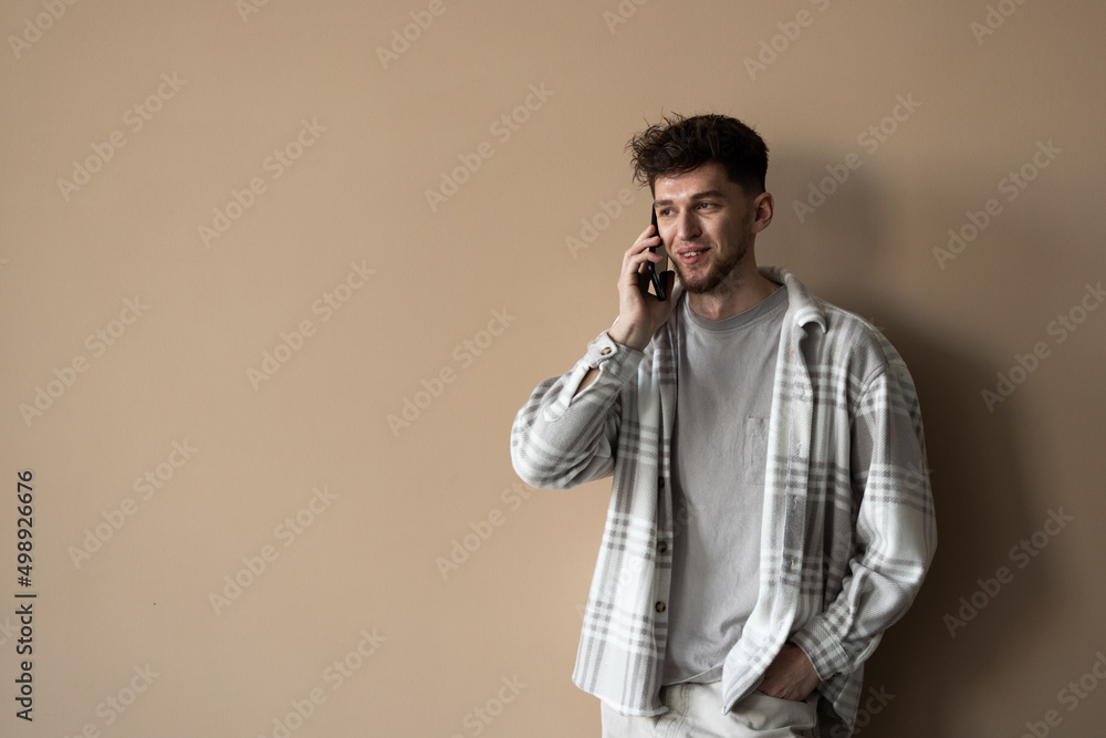 Young man wearing shirt smiling and talking on mobile phone isolated over beige background Young man wearing shirt smiling and talking on mobile phone isolated over beige background