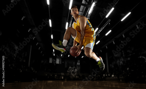 In action. Young basketball player jumping with ball in flashlights over dark gym background. Concept of sport, energy and dynamic, healthy lifestyle. Arena's drawned