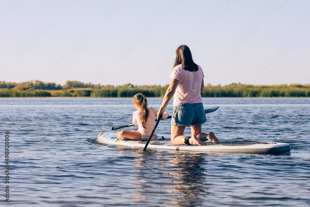 Mother and little daughter on sup board rowing together effortfully, exercising and having fun on lake with green reeds in background. Active lifestyle. Teaching to do sports from childhood.