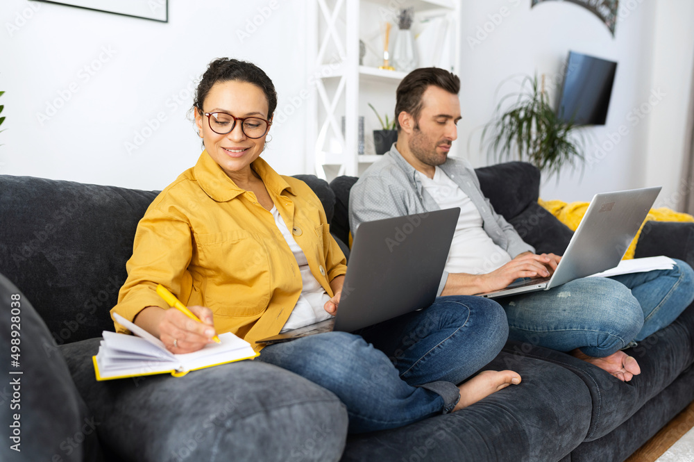 Couple using laptops for remote work on the sofa at home, focused spouses working on the distance, answering emails, doing paperworks in home atmosphere