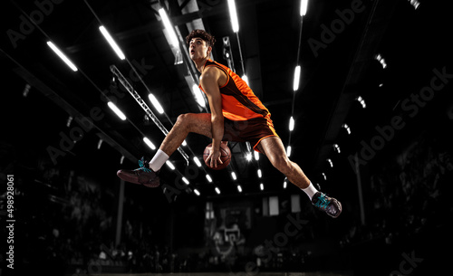 In action. Young basketball player jumping with ball in flashlights over dark gym background. Concept of sport, energy and dynamic, healthy lifestyle. Arena's drawned