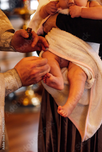 Fotografia Cropped photo of priest in cassock hold bottle with holy oil for anointing feet of baby in white towel holded by godmother during ceremony of infant baptism in orthodox church