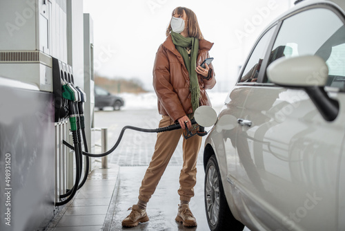 Woman in face mask refueling car with a gasoline, using smartphone to pay. Concept of mobile technology for fast refueling without visiting the store photo