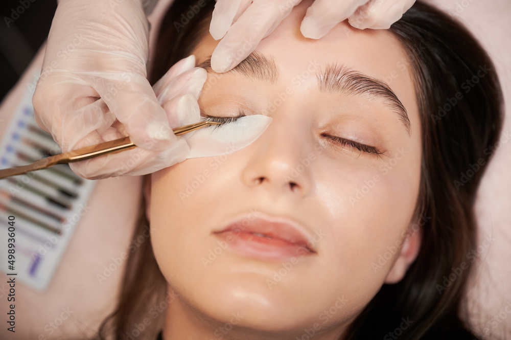 Close up of lash specialist in sterile gloves applying lash extensions with tweezers. Young woman with eye patch under lower eyelid keeping eyes closed while having eyelash extension procedure.