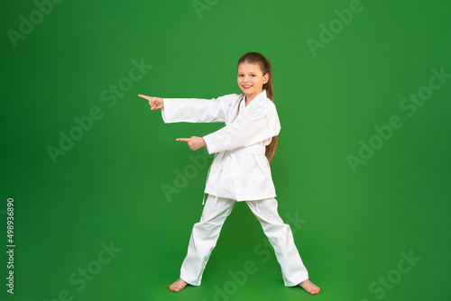 A girl in a kimono on a green background points her fingers at the advertisement. kid studies martial arts. child development in sports. © Юлия Дьякова