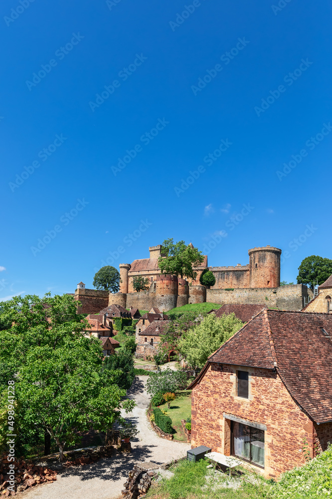 Road leading through small village and lush greenery to hilltop fortress Chateau de Castelnau-Bretenoux. Prudhomat, Lot, Occitanie, France