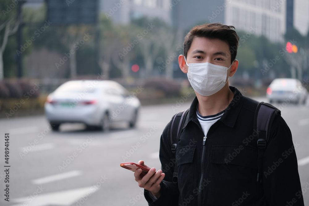 Asian young man in face mask holding phone to hail taxi near road, looking at camera Asian young man in face mask holding phone to hail taxi near road, looking at camera