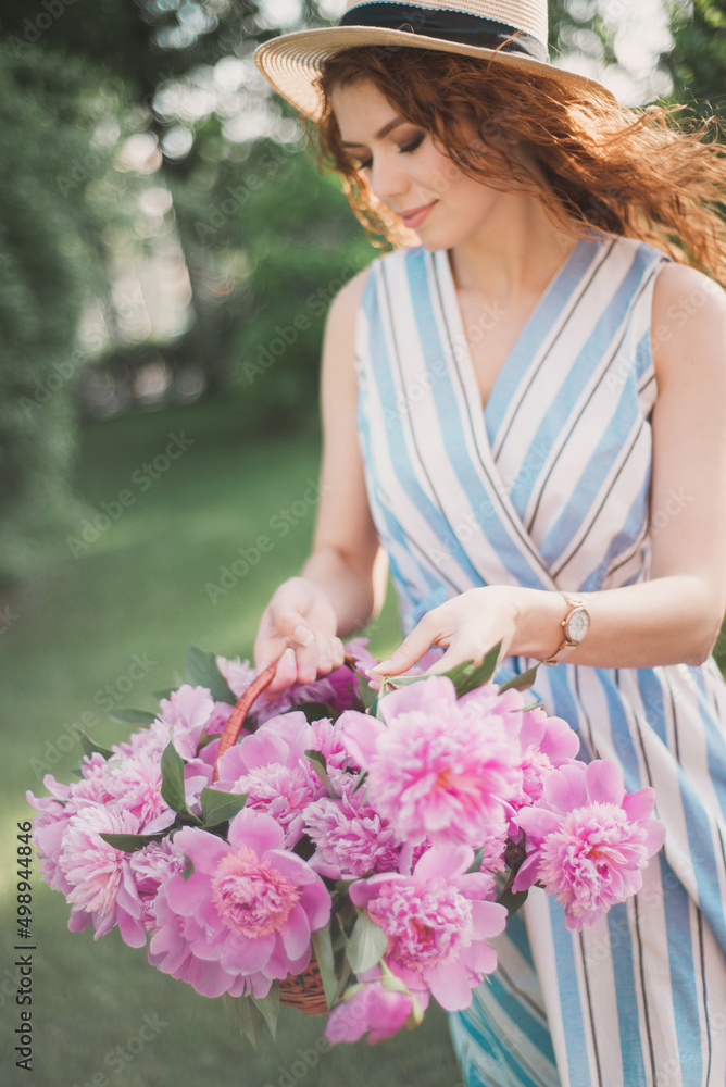 Portrait of young redhead curly woman in straw hat and linen stripe dress with a basket and a pink  peonies bouquet in the garden. No face closeup