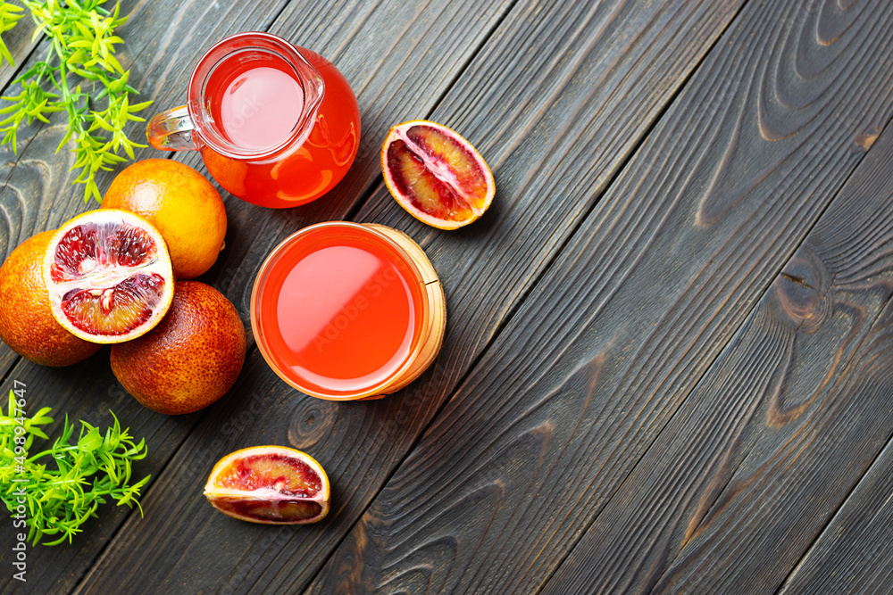 A transparent glass with red orange juice on a wooden table. Red oranges and green twigs in the background. Wooden background, space for text A transparent glass with red orange juice on a wooden table. Red oranges and green twigs in the background. Wooden background, space for text