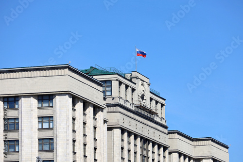 Parliament building in Moscow with Russian flag on background of clear blue sky. Facade of State Duma of Russia with soviet coat of arms, russian authority