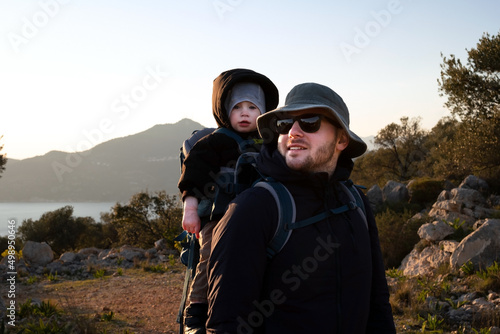 Man wearing sunhat, sunglasses carrying baby,toddler in backpack.Young handsome father traveling,hiking with kid, child of 1,2 years old.Family trip in nature,mountains,cold weather near sea