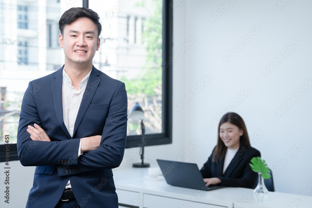 Asian young successful businessman wear formal suit smiling with confident and standing with crossed arm at office with his colleagues woman working on laptop on blur background. White background Asian young successful businessman wear formal suit smiling with confident and standing with crossed arm at office with his colleagues woman working on laptop on blur background. White background