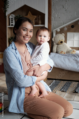 Portrait of loving young mother wearing casual clothes sitting on floor holding baby in arms smiling at camera