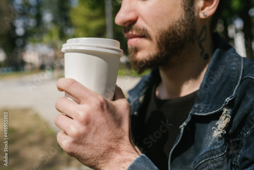 Close up view of young man drinking coffee. He is wearing jean jacket and black sweater. He looks away and thinks.