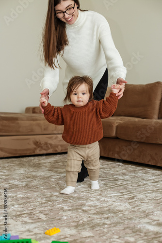 Cute little girl takes first steps with help of her mother, home interior, close up. Happy childhood photo