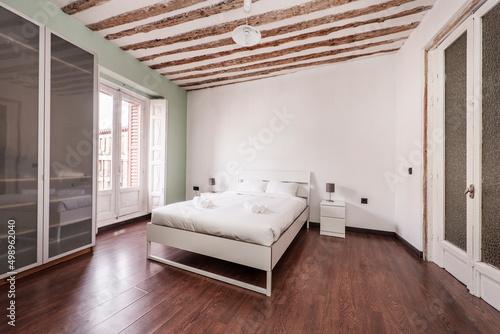 Bedroom with large bed with white sheets  clean white towels  wardrobe with glass doors  exposed wooden beams on the ceiling and balcony with white wooden shutters