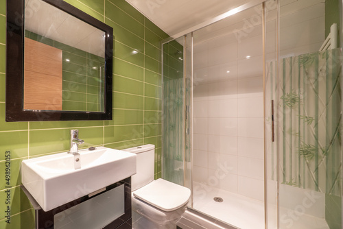Bathroom with green tiles, wall mirror with wooden frame, white porcelain sink on brown wooden cabinet and shower cabin with glass screen and water jets