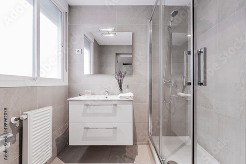 Bathroom with aluminum window, frameless wall mirror, white porcelain sink on white marble top and shower cabin with glass screen and water jets